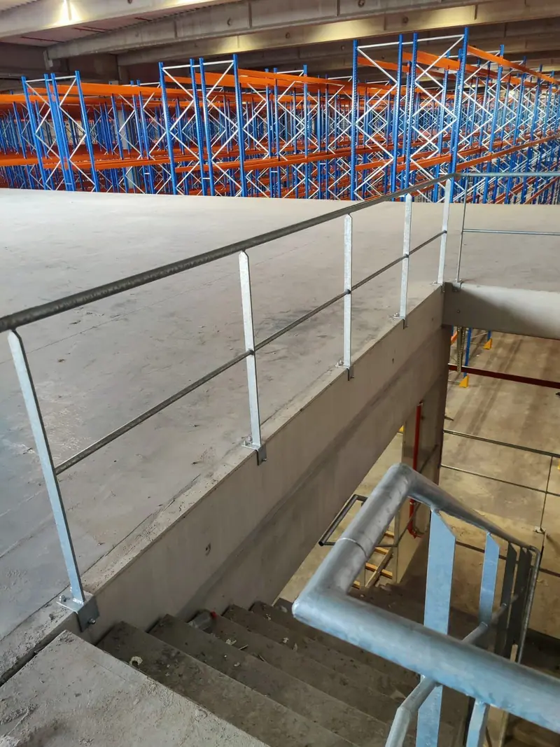 Stalowe barierki z rur na krawędzi antresoli i przy schodach w hali z regałami magazynowymi