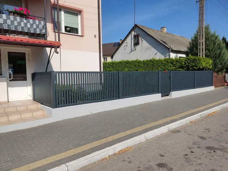 Ogrodzenie z pionowych profili stalowych z furtką, na podmurówce wzdłuż chodnika przy budynku